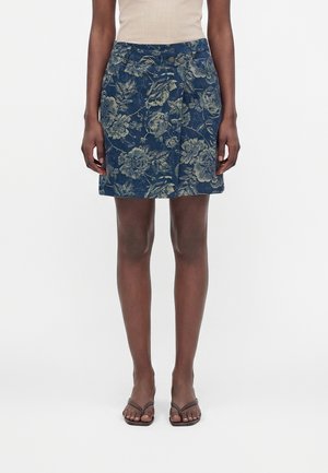 Kvinde iført en knælængde blå nederdel med beige blomsterprint, beige ærmeløs top og mørkebrune flip-flop sandaler, stående mod hvid baggrund.