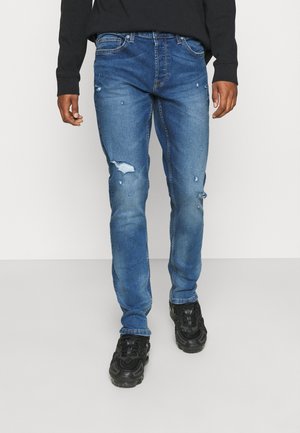 Mand iført blå slidte jeans, sorte sneakers og en sort langærmet top, stående mod en enkel hvid baggrund.