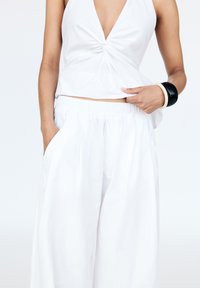 Weißes ärmelloses Oberteil mit einem geknoteten Design in der Mitte, kombiniert mit lockeren weißen Hosen mit seitlichen Taschen und einem elastischen Bund.