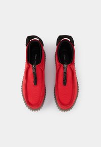 Chaussures rouges à enfiler en matériau synthétique, avec une fermeture éclair noire et des semelles en caoutchouc à adhérence texturée. Accents noirs autour du col.