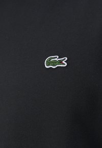 Tissu en coton noir avec un petit logo crocodile vert brodé, orné d'un accent rouge, placé près du coin supérieur gauche.