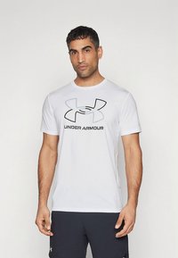 Bela bombažna majica kratkih rokavov z velikim črnim in sivim logotipom Under Armour na prsih, z okroglim izrezom in sproščenim krojem.