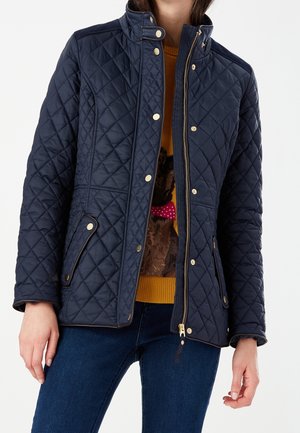 Kobieta w granatowej pikowanej kurtce z złotymi guzikami, częściowo rozpięta, na musztardowym swetrze z grafiką psa i ciemnoniebieskich dżinsach.