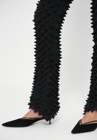 Musta tekstuuriga püksid, millel on terava disain, mis ulatub mööda jalgu allapoole, koos terava nokaga mustade sametkingadega, millel on peen vähese lõike detail.