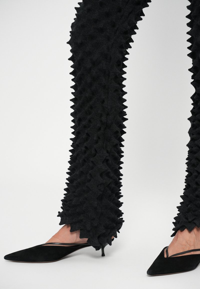 Pantaloni neri in tessuto testurizzato con un design a punte che si estende lungo le gambe, abbinati a tacchi a punta neri in suede con un delicato dettaglio a taglio.