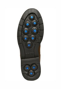 Semelle noire avec des accents circulaires bleus et des trous de ventilation, texturée pour l'adhérence. Présente une partie supérieure en suede marron clair.