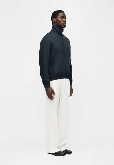 Homme debout regardant vers la droite, portant une veste sombre à col montant, un pantalon blanc à jambes larges et des chaussures noires sur un fond clair uni.