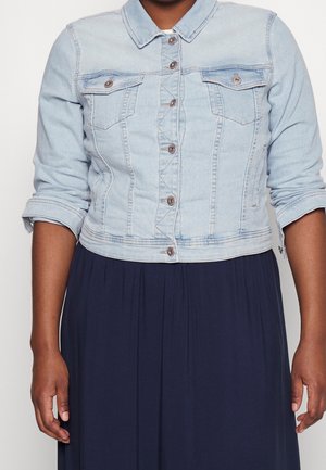 Vrouw draagt een lichtblauwe cropped spijkerjas met opgerolde mouwen over een donkerblauwe geplooide jurk, weergegeven van nek tot heupen.