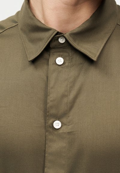 Γιακάς και άνω μέρος του στήθους ενός πουκαμίσου ελιάς με κουμπιά, με τρία λευκά κουμπιά κουμπωμένα, που φοριέται από άτομο με ανοιχτόχρωμο τόνο δέρματος.