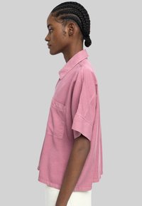 Koszulka z krótkim rękawem o luźnym kroju w kolorze przybrudzonego różu, wykonana z bawełny. Posiada kołnierzyk oraz kieszeń na piersi, charakteryzuje się miękką fakturą i bocznymi rozcięciami.