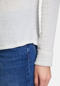 White textured long-sleeve shirt with a curved hem, paired with blue denim. Tag visible near the bottom hem on the left side.