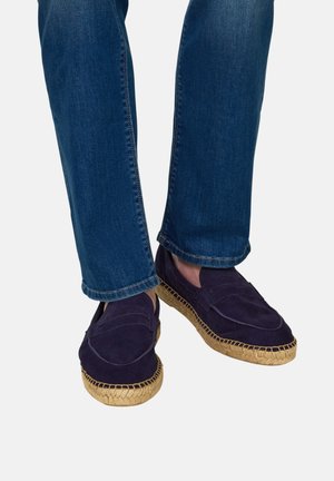 Persoon die donkerblauwe suède espadrille loafers met natuurlijke geweven zolen en blauwe spijkerbroek draagt, staand op een effen achtergrond.