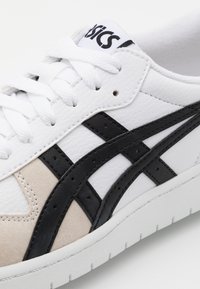 Baskets en cuir blanc avec des accents noir et beige, présentant un design bas, des lacets plats et une semelle en caoutchouc texturée.