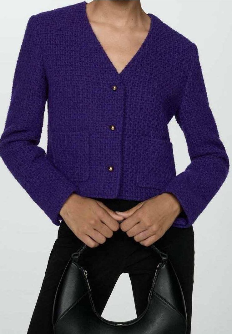 Femme portant une veste violette texturée boutonnée, tenant un sac à main en cuir noir à deux mains devant la taille.