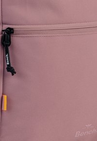 Roze textuur van de rugzak met een horizontale rits, met een zwarte trekker met het "Bench"-logo en een klein geel accent aan de zijkant.
