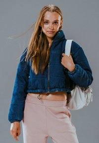 Krótka niebieska kurtka z zamkiem błyskawicznym i elastycznymi mankietami, zestawiona z różowymi spodniami dresowymi i białym plecakiem, na szarym tle.