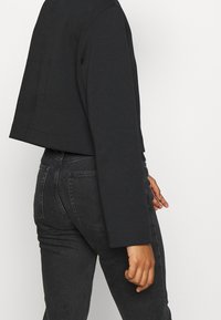 Svart kort jacka med långa ärmar, strukturerat tyg och rak fåll, kombinerad med höga svarta jeans i denim.