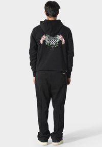 Uomo che indossa un hoodie nero e pantaloni, con la schiena che mostra una grafica a tema corsa con bandiere a scacchi e il testo "Pas De Monaco".