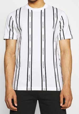 T-shirt blanc à manches courtes avec des rayures noires verticales et le texte "Calvin Klein" répété, porté par une personne les bras le long du corps.