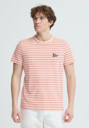 Junger Mann, der ein hellrosa T-Shirt mit dünnen roten horizontalen Streifen und weiße Hosen trägt, schaut leicht nach links vor einem schlichten Hintergrund.