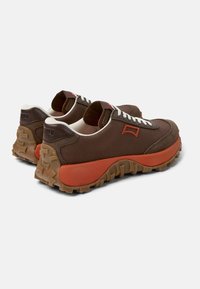Sneakers in tessuto marrone con lati testurizzati, suola intermedia arancione e suola robusta. Presentano lacci bianchi e un piccolo logo accento di lato.