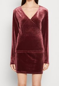 Kobieta w długim rękawie, sukience wrap V-neck z burgundowego weluru, z krótkim dołem, stojąca na tle gładkiej, jasnej ściany.
