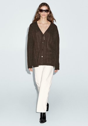 Cardigan marrone in maglia texturizzata con scollo a V profondo, chiusura a bottoni, indossato sopra pantaloni bianchi a gamba larga, completato da stivaletti neri e occhiali da sole.