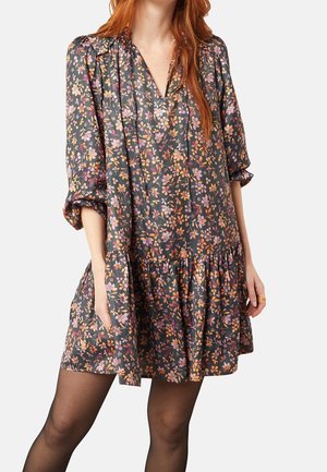 Mujer con un vestido floral suelto con flores rosas, naranjas y moradas sobre medias negras transparentes, sobre un fondo liso.