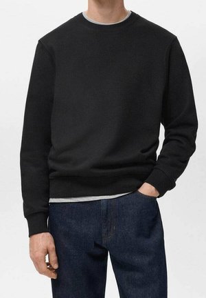 Mand iført en sort langærmet crewneck-trøje, mørkeblå jeans og en grå skjorte under, med den ene hånd i lommen.