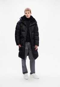 Sort puffede jakke med en pelsforet hætte, lynlås, sidelommer og en puffy design. Bæres over en sort rullekrave og grå bukser.