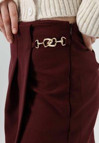 Bordeauxrode broek met een gouden kettingaccent bij de taille, voorzien van een zijrits en geplooide details in de stof. Glad aanvoelend.