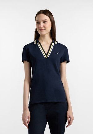Kvinde iført en marineblå kortærmet polo med hvide, gule og mørke stribeaccenter på kraven mod en ensfarvet baggrund.