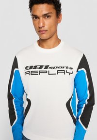 White long-sleeve shirt featuring black and blue accents, with "981sports REPLAY" printed in bold black text across the front.