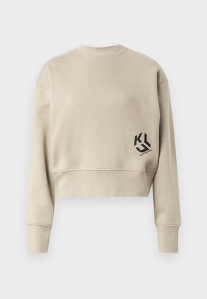 Afkortet sweatshirt i lys beige stof. Har lange ærmer, rund halsudskæring, og et sort logo trykt på nederste venstre side.