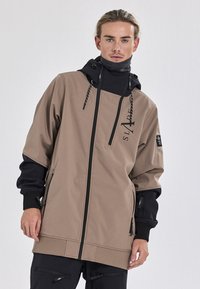 Beige und schwarze Kapuzenjacke mit Reißverschluss, ausgestattet mit einem Logo, verstellbaren Kordeln und Reißverschlusstaschen an den Ärmeln. Glatte Textur.