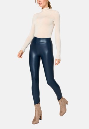Frau trägt einen cremefarbenen Rollkragenpullover, marineblaue Leggings aus Kunstleder und beige Wildleder-Stiefeletten, läuft vor neutralem Hintergrund.
