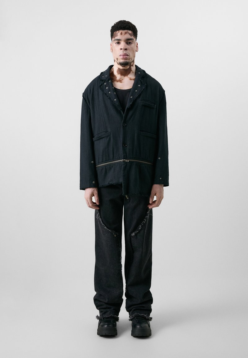 Schwarze, oversized Jacke mit fransigen Kanten, zwei Taschen und Knopfverschluss. Kombiniert mit ausgefransten schwarzen Hosen und klobigen schwarzen Stiefeln.