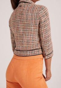 Rakenteellinen, monivärinen tweed-takki, jossa on kaulus, erottuva musta viimeistely ja kolmasosa hihat. Yhdistetty appelsiininkeltaisiin korkeavyötäröisiin housuihin.