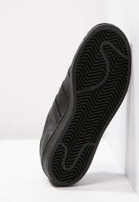 Baskets noires avec une semelle en caoutchouc texturée, présentant un motif ondulé et trois bandes latérales. Partie supérieure lisse avec un design élégant.