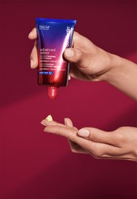 Crema idratante al retinolo in un tubo blu e rosso con punta appuntita; una piccola quantità di prodotto cremoso, giallo pallido su una mano. Lo sfondo è di un rosa scuro.