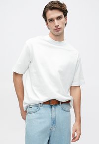 Mladý muž s krátkymi hnedými vlasmi, oblečený v obyčajnom bielom tričku zasunutom do svetlomodrých džínsov s hnedým opaskom, stojí pred bielym pozadím.