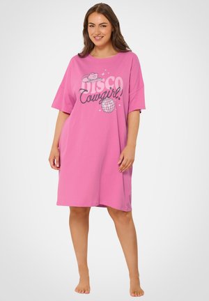 Smilende kvinde iført løs lyserød kjole med teksten "Disco Cowgirl", cowboyhat og diskokuglegrafik, stående barfodet på hvid baggrund.