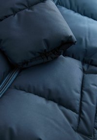 Primo piano di una giacca a piumino blu navy, caratterizzata da un design trapuntato, una texture imbottita e una cerniera visibile con linguetta abbinata.