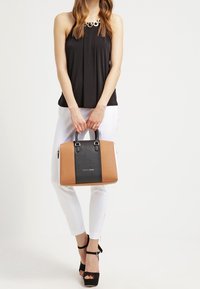 Borsa strutturata in pelle nera e marrone chiaro, forma rettangolare, manici doppi e dettagli metallici. Il modello indossa una maglietta nera e dei pantaloni bianchi.