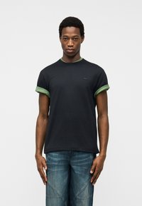 Νεαρός άνδρας όρθιος, φοράει μαύρο μπλουζάκι με πράσινες διπλωμένες μανσέτες και μπλε τζιν, μπροστά σε απλό ανοιχτό γκρι φόντο.