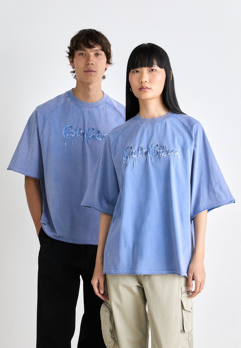 Två individer bär överdimensionerade blå t-shirts med ett droppande textdesign. Tyget verkar mjukt; tröjorna har korta ärmar och rund halsringning.