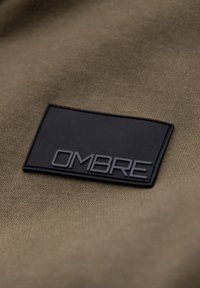 Patch rectangulaire noir avec le mot "OMBRE" en relief, placé sur un fond en tissu texturé vert olive.
