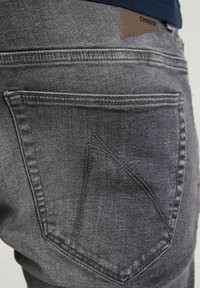 Primer plano del bolsillo trasero de unos jeans de mezclilla gris con detalles de costura y una etiqueta de marca marrón que dice "CHASIN'" cerca de la cinturilla.