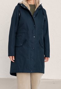 Granatowy nieprzemakalny płaszcz z kapturem, zatrzaskami i dwoma przednimi kieszeniami. Gładka faktura, długość do połowy uda i regulowany sznurek.