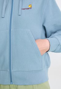 Sudadera con cremallera de color azul claro, hecha de un tejido suave. Presenta un bolsillo tipo canguro y puños acanalados. Logo en amarillo y marrón en el pecho.